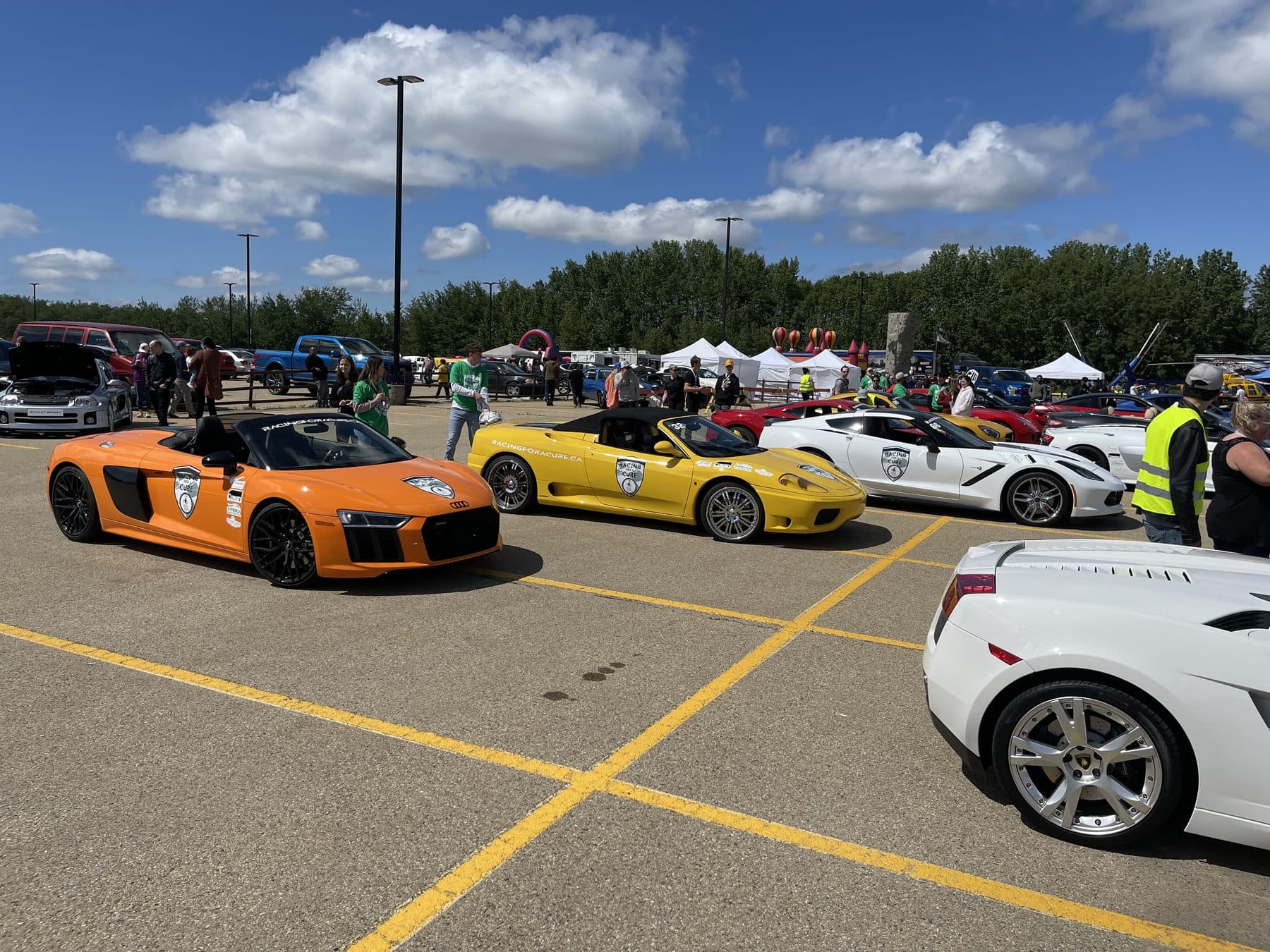 A lineup of supercars — an orange Audi R8, a yellow Ferrari and a white Lamborghini — in a parking lot before a Racing for a Cure event.