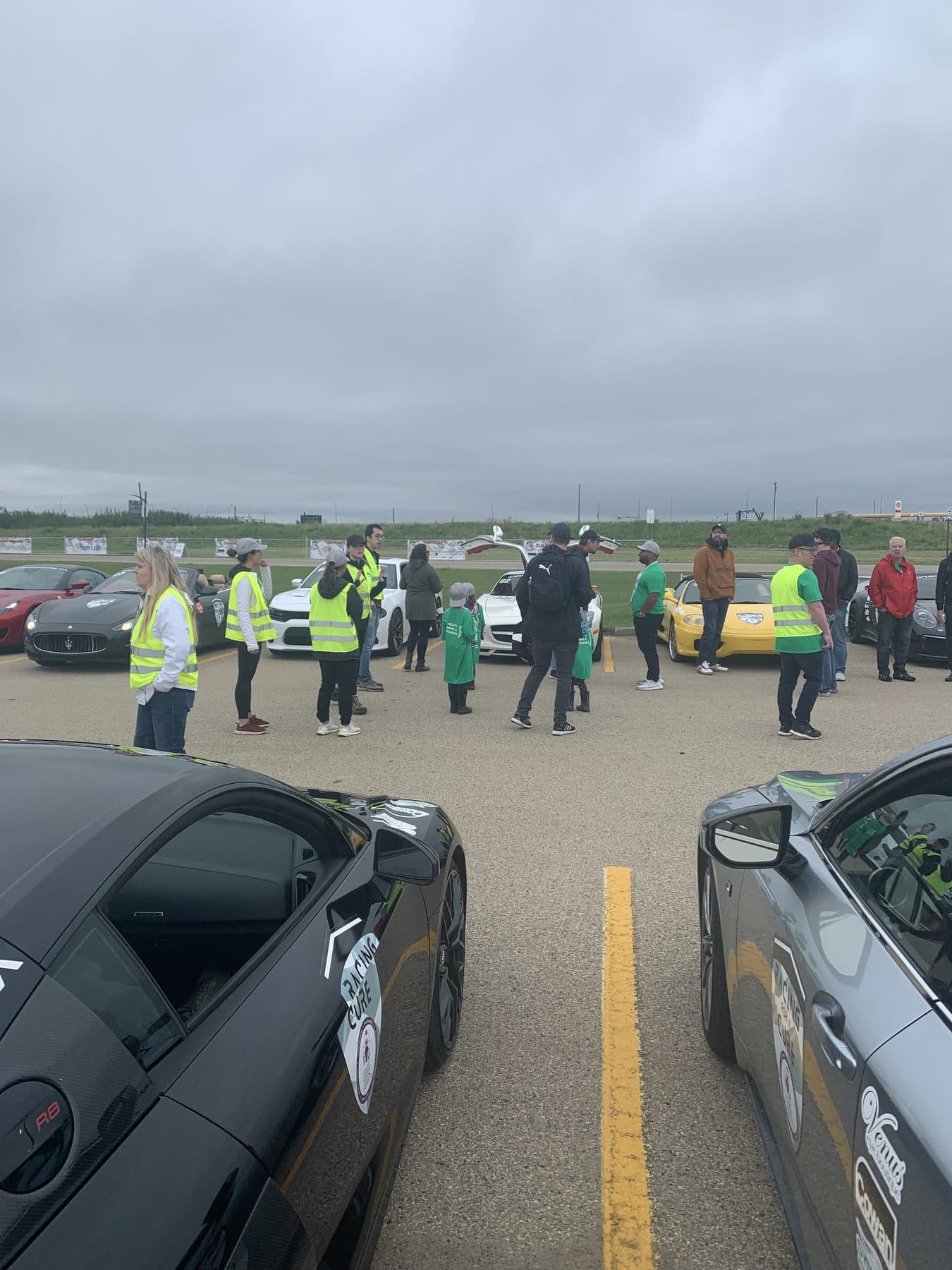 Volunteers in green shirts and high-visibility vests gathering between sports cars at a charity racing event.